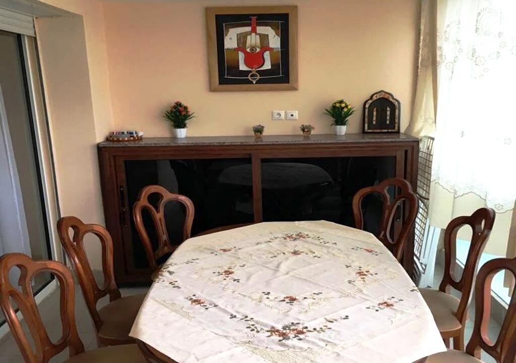A dining area with a round table covered with a floral tablecloth, surrounded by wooden chairs. There is a wooden cabinet with decorative items on top and a framed artwork above it.