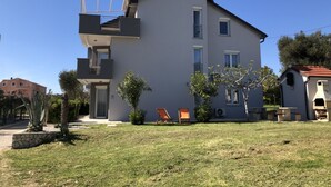 Un bâtiment moderne à deux étages, doté d’un toit plat, d’un balcon à l’étage supérieur et d’un petit jardin avec une pelouse et quelques plantes en pot.