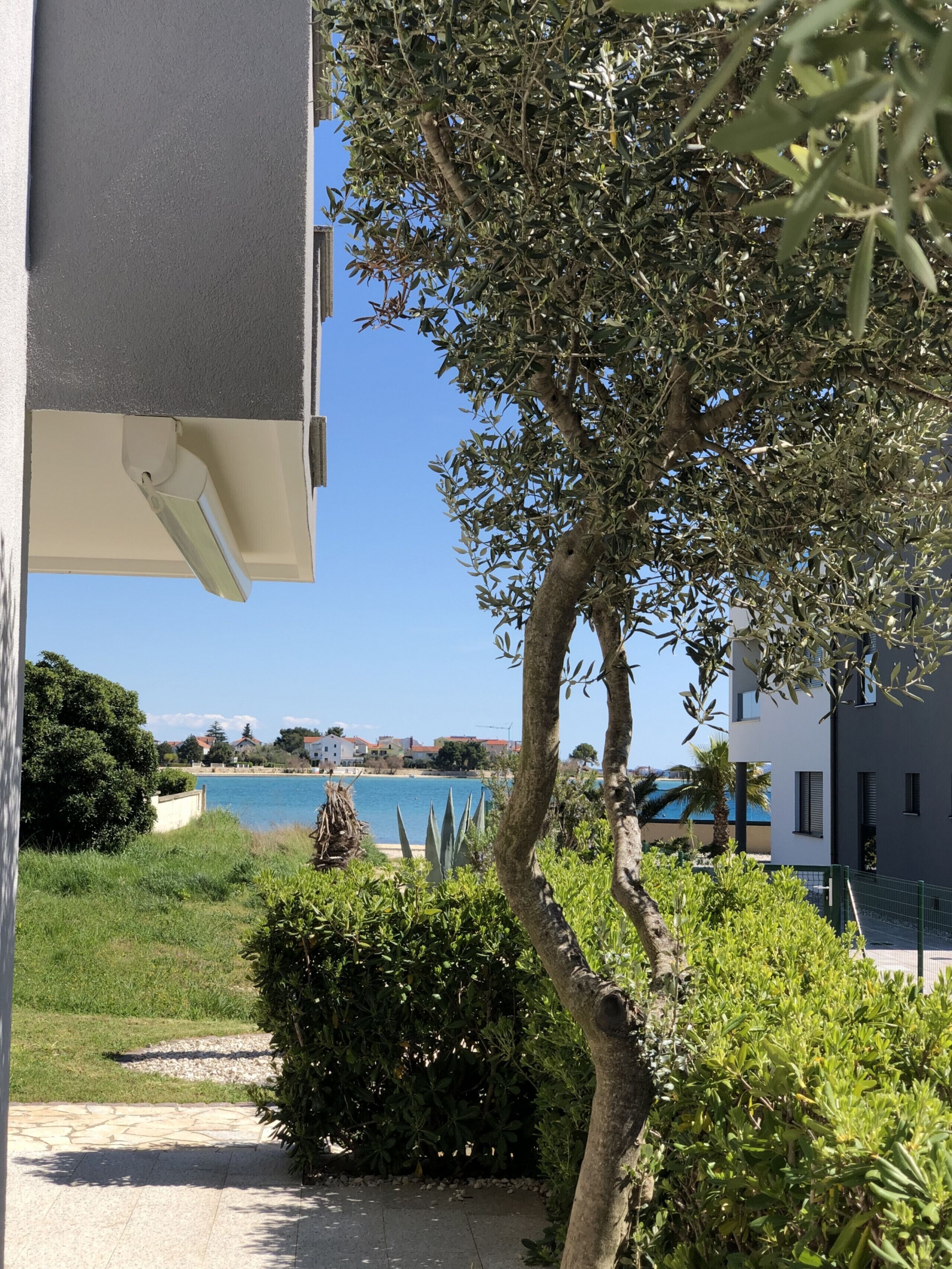 A view of a coastal area with a clear blue sea, a pathway leading to a building, and lush greenery.