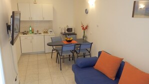Un espace de vie compact comprenant un coin cuisine, une table à manger avec des chaises et un canapé bleu agrémenté de coussins orange.