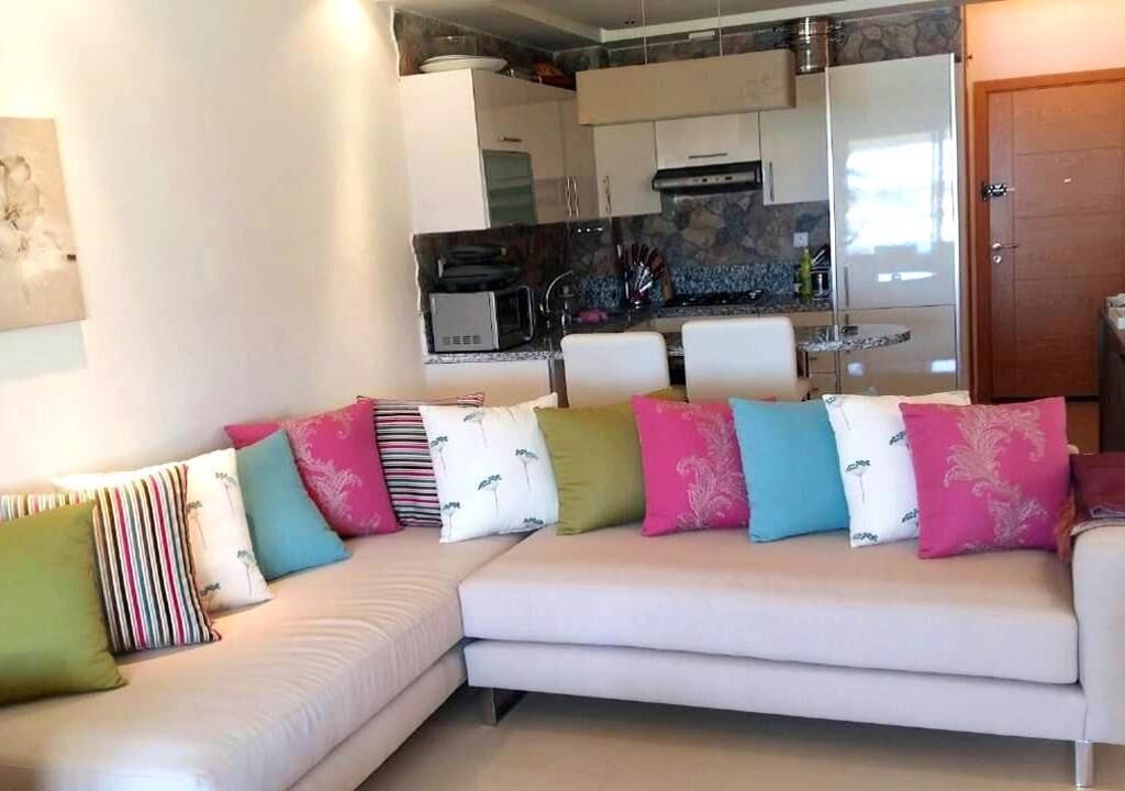 Un salón moderno con un sofá blanco, cojines de colores, una cocina al fondo y una puerta de madera.