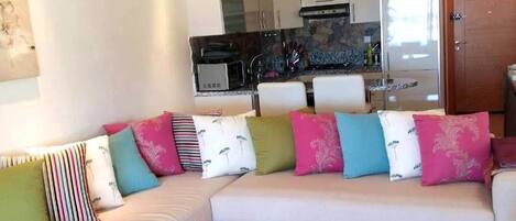Un salón moderno con un sofá blanco, cojines de colores, una cocina al fondo y una puerta de madera.