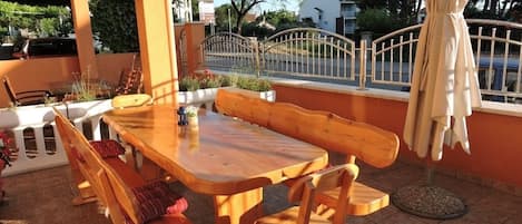 En udendørs terrasse med et træbord og bænke, omkranset af et hvidt hegn og udsigt til en gade og huse.
