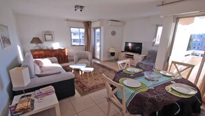 A living room with a sofa, a coffee table, a dining table set for two, a TV, and a balcony with a view.