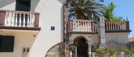 A building with a stone facade, a balcony with white balustrades, and a gate with a number '165' above it.