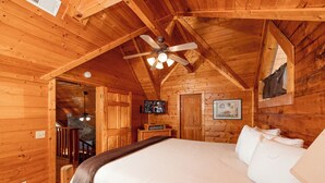 Ein Schlafzimmer in einer Blockhütte mit Bett, Deckenventilator, Fernseher und einer Wand aus Stein.