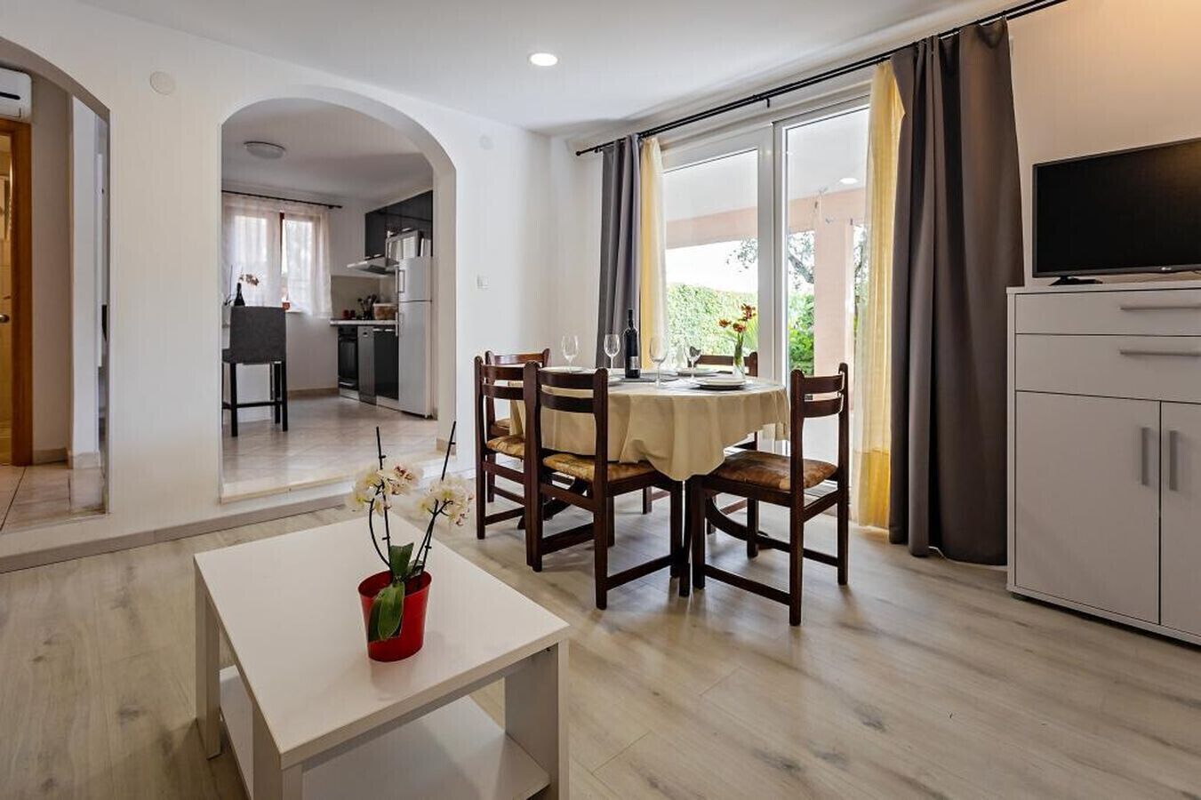 A modern living room with a dining area, a kitchen in the background, and a TV on a cabinet.