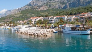 A coastal marina with boats docked, a rocky shoreline, and buildings with red-tiled roofs in the background. - Apartment in Tučepi with Terrace, Air condition, WIFI, Washing machine (202-3) (Tucepi)