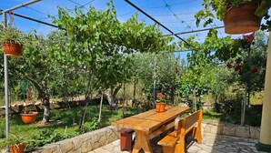 Um pátio externo com mesa e bancos de madeira, rodeado por um muro de pedra e vegetação exuberante.