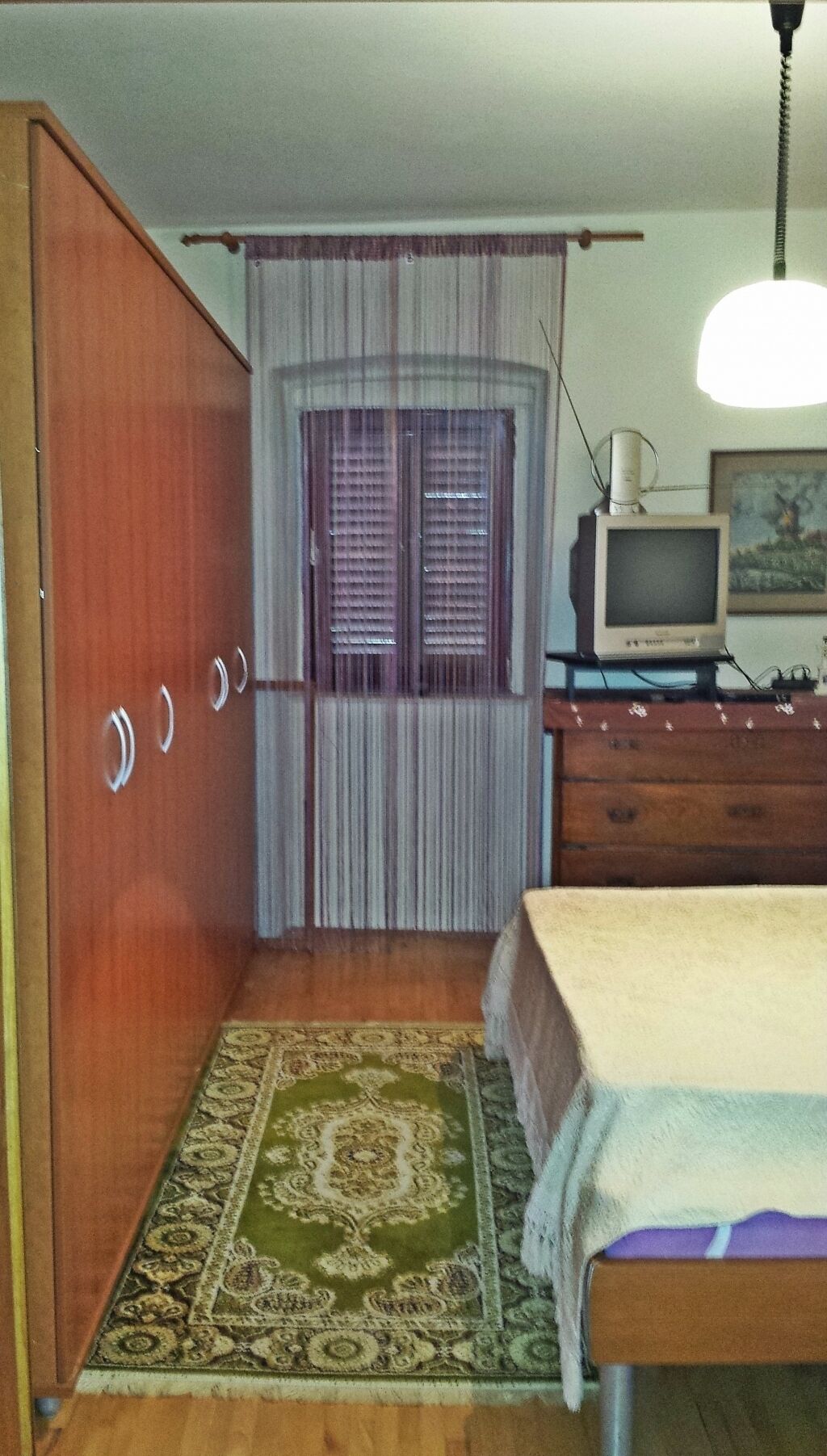 A room with a wooden wardrobe, a bed with a white cover, a television on a wooden dresser, and a patterned rug on the floor.