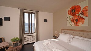 Une chambre d’hôtel avec un lit, une fenêtre avec des rideaux, une vue sur la mer, un tableau représentant des fleurs rouges et une petite table avec une plante.