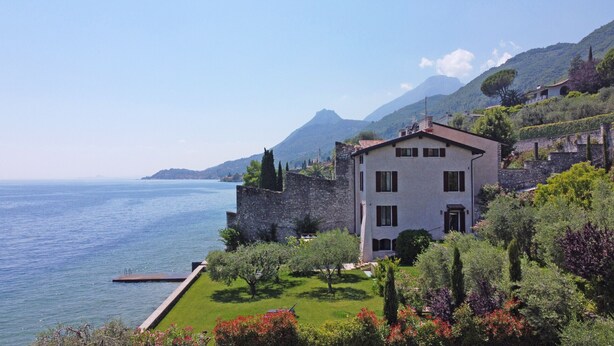 Un bâtiment à deux étages, doté d’un mur en pierre et d’un jardin à l’avant, qui offre une vue sur une étendue d’eau et des montagnes.