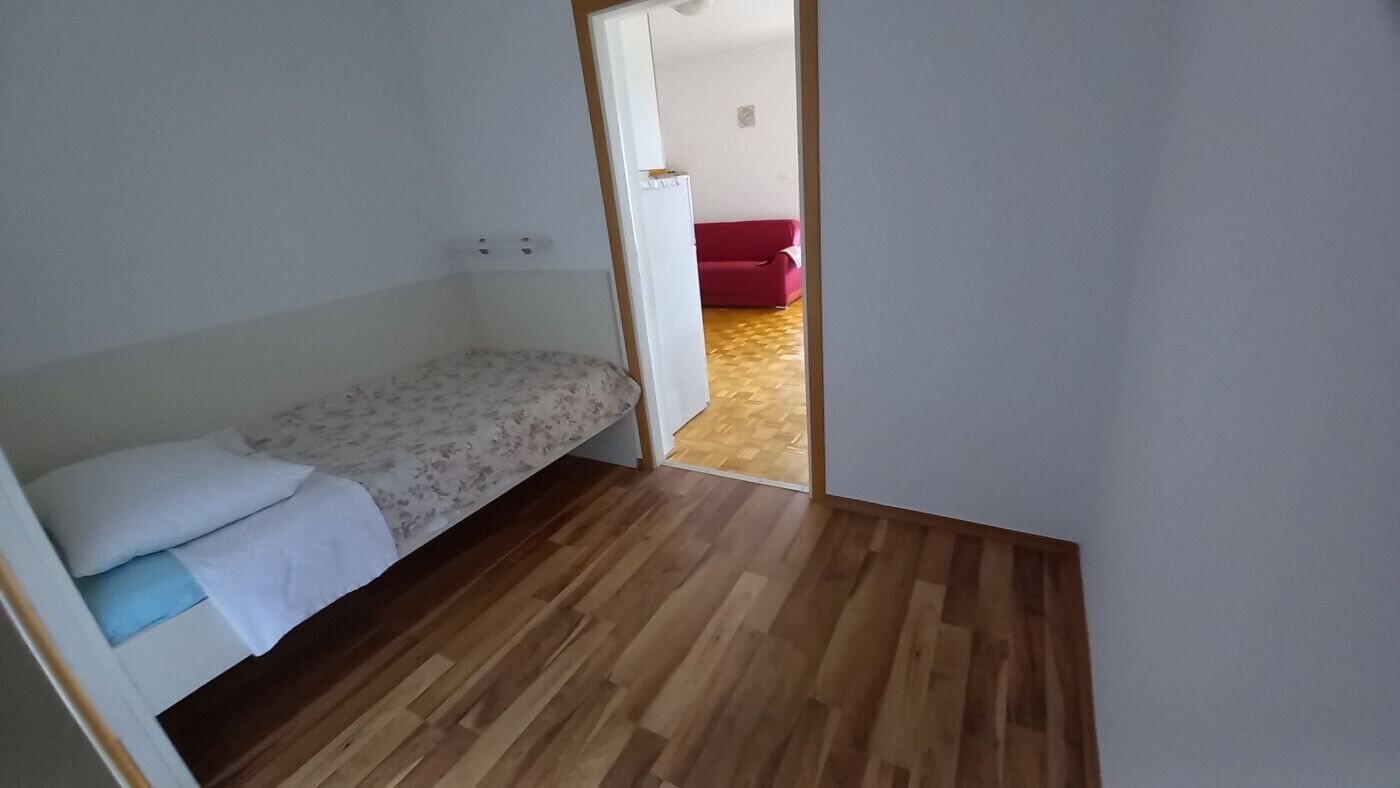 Una stanza piccola con pavimento in legno, un letto con copriletto floreale e un divano rosso sullo sfondo.