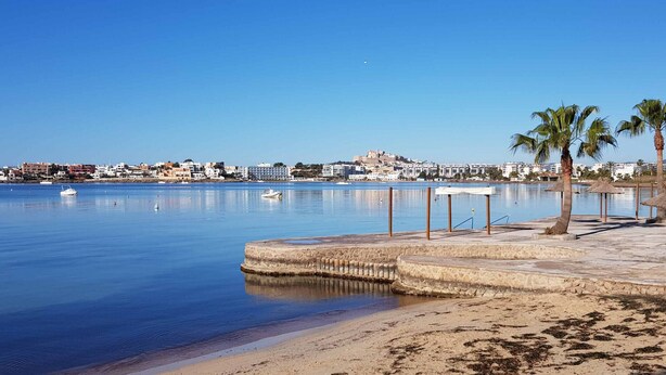 Una orilla tranquila con playa de arena, palmeras y un pueblo lejano.