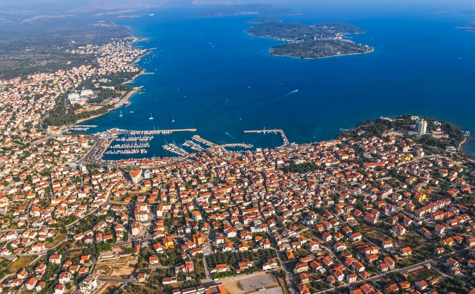 Luftaufnahme einer Küstenstadt mit dichter Bebauung, einem Yachthafen und einer markanten Insel.