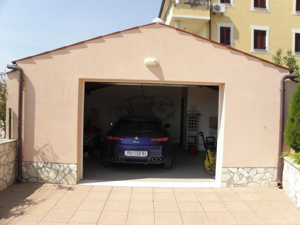 Un garage dans lequel est garée une voiture bleue, un mur de pierres et, au fond, un bâtiment jaune.