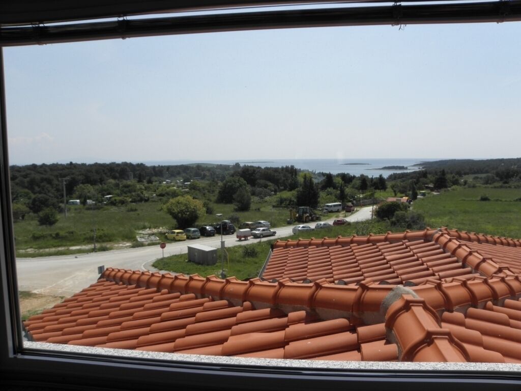 Blick aus einem Fenster auf ein terrakotta-gefliesstes Dach, einen Parkplatz und eine entfernte Landschaft mit Bäumen und einem Gewässer.