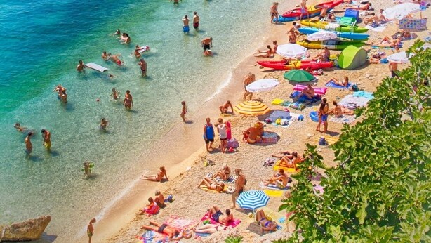 Une plage avec une eau turquoise et limpide, des gens qui se baignent et prennent des bains de soleil, et un bâtiment historique en arrière-plan.