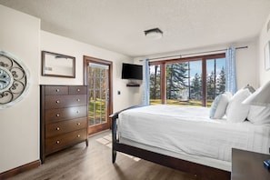 A bedroom with a bed, a dresser, a television, and a window with a view of trees.