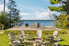 Une terrasse dotée de chaises Adirondack donnant sur un lac, avec un foyer et une rambarde offrant une vue sur l’eau.