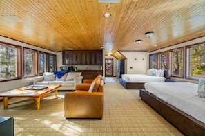 A spacious room with a wooden ceiling, large windows, a sofa, a bed, and a kitchen area. - The Stone House  | Premier West Twin Lake Home (Lewiston)