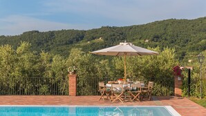 A poolside area with a table and chairs, an umbrella, and a view of a forested hill.