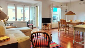 Una sala de estar con un sofá, una silla de madera, una mesa de comedor con sillas, un televisor y un ventanal grande con persianas.