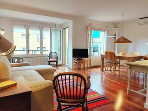 Un salon comprenant un canapé, une chaise en bois, une table à manger avec des chaises, un téléviseur et une grande fenêtre avec des stores.