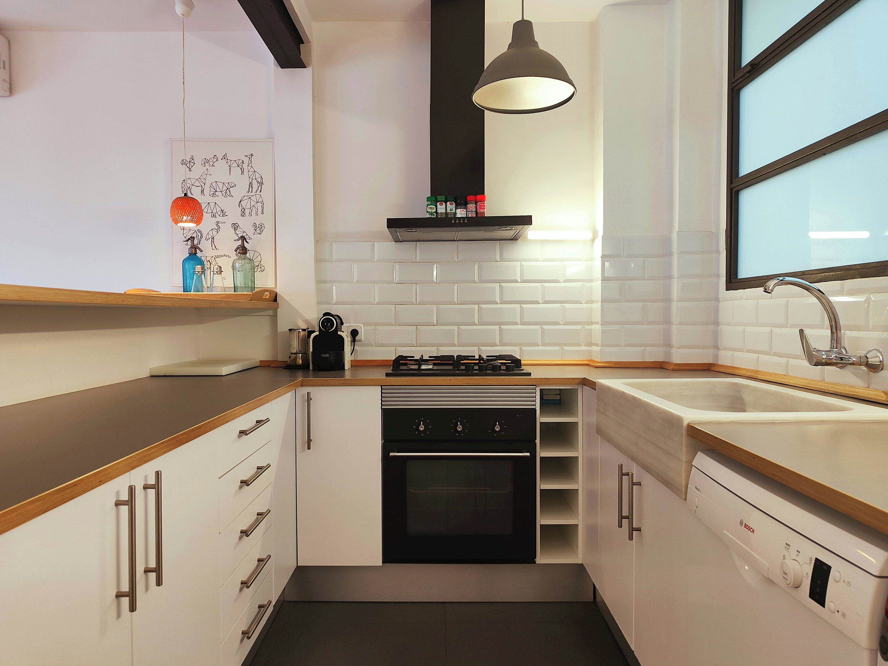 Une cuisine compacte avec des meubles blancs, un four noir, un évier en acier inoxydable et un luminaire suspendu.