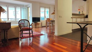 Amplia sala de estar con piso de madera, una mesa de comedor, un televisor y una cocina con armarios blancos.
