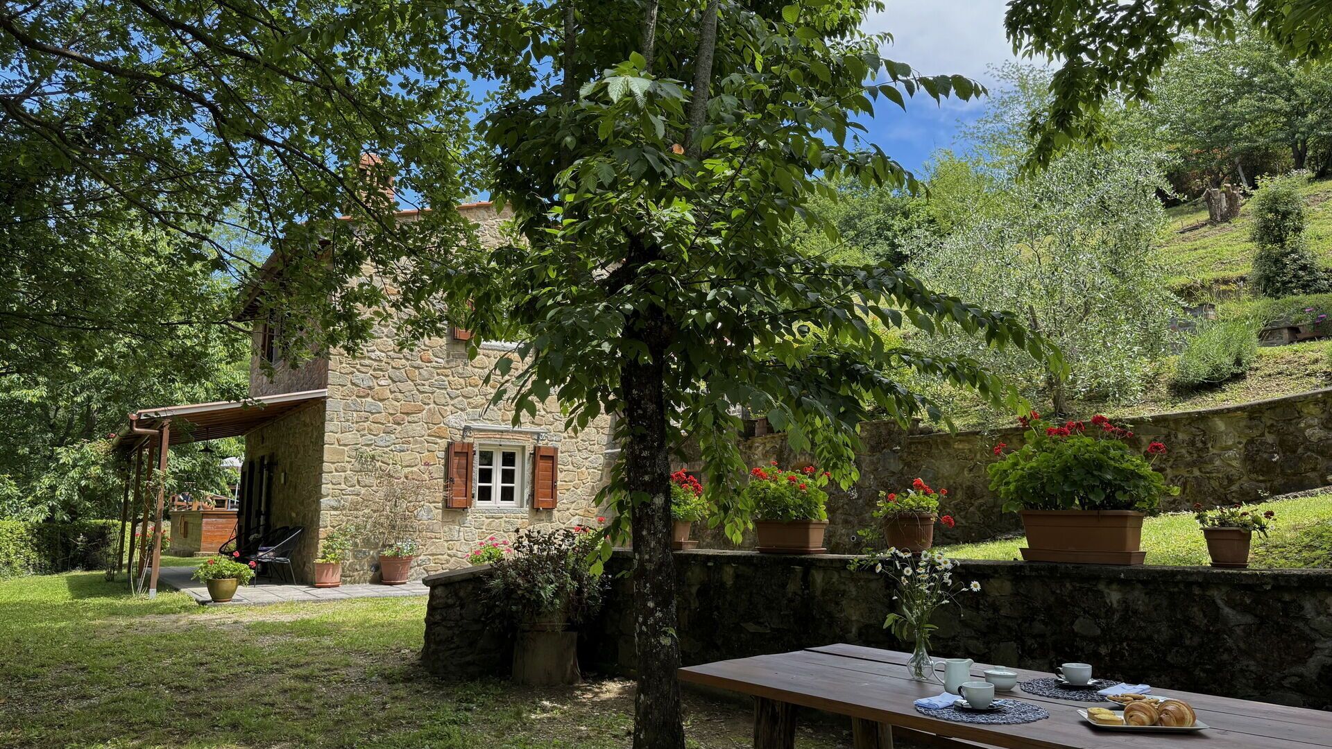 Ein Steinhaus mit einer Holzveranda, umgeben von üppiger Vegetation und Pflanzgefäßen.