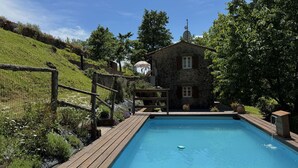 Ein Schwimmbad mit Holzterrasse, umgeben von einem Steinhaus und üppiger Vegetation.