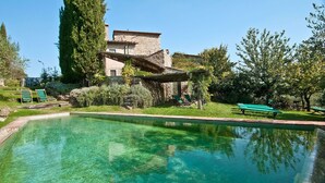 Une piscine aux eaux cristallines, avec un bâtiment en pierre en arrière-plan, entourée de verdure et agrémentée d’un banc.