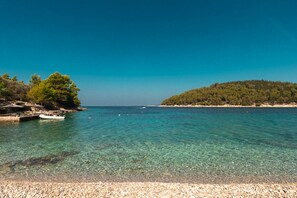 A clear coastal scene with a pebble beach, a small boat docked near the shore, and a forested island in the distance.