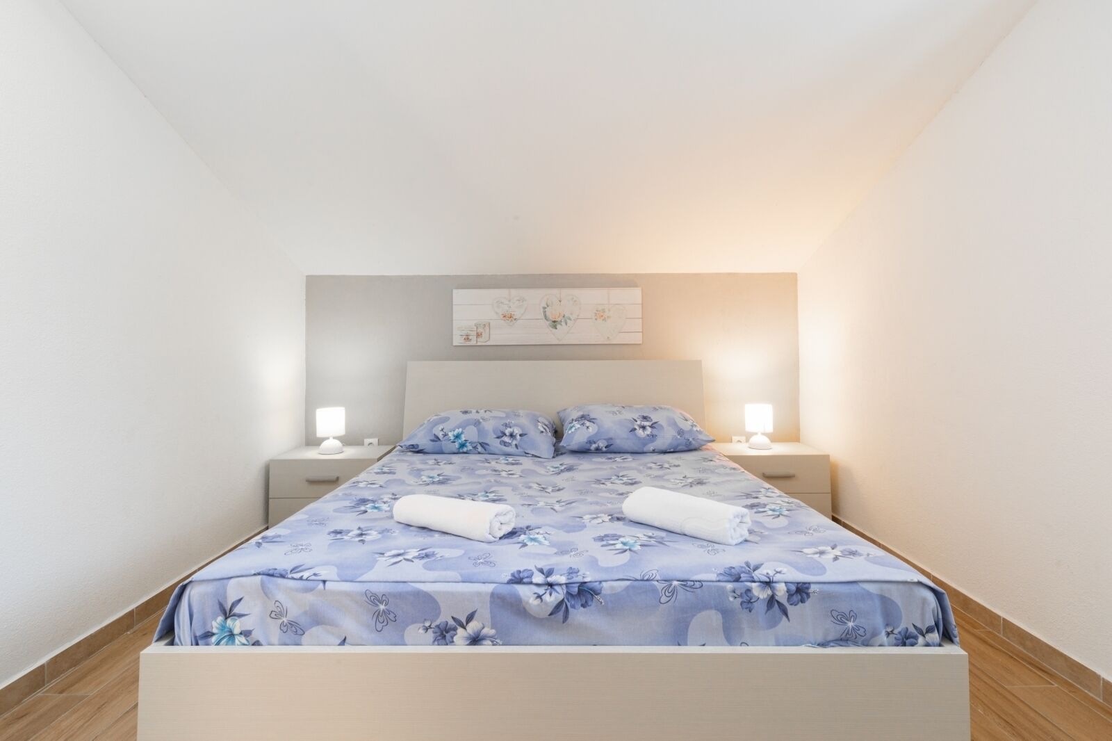 Ein ordentlich bezogenes Bett mit floralem Bettbezug und zwei aufgerollten Handtüchern, ein Nachttisch mit Lampe und ein an der Wand befestigtes Kunstwerk.