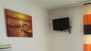 Un lit simple avec du linge de lit blanc, un téléviseur fixé au mur et un tableau représentant un coucher de soleil sur l’eau.