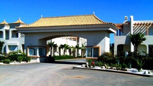A large building with a prominent entrance under a tiled roof, surrounded by landscaped gardens and palm trees. - Nice apartment in Mozona Beach 4205 (Sidi Rahal)