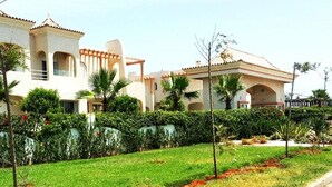 A residential area with a well-maintained lawn, a row of palm trees, and a building with a tiled roof and arches. - Nice apartment in Mozona Beach 4205 (Sidi Rahal)