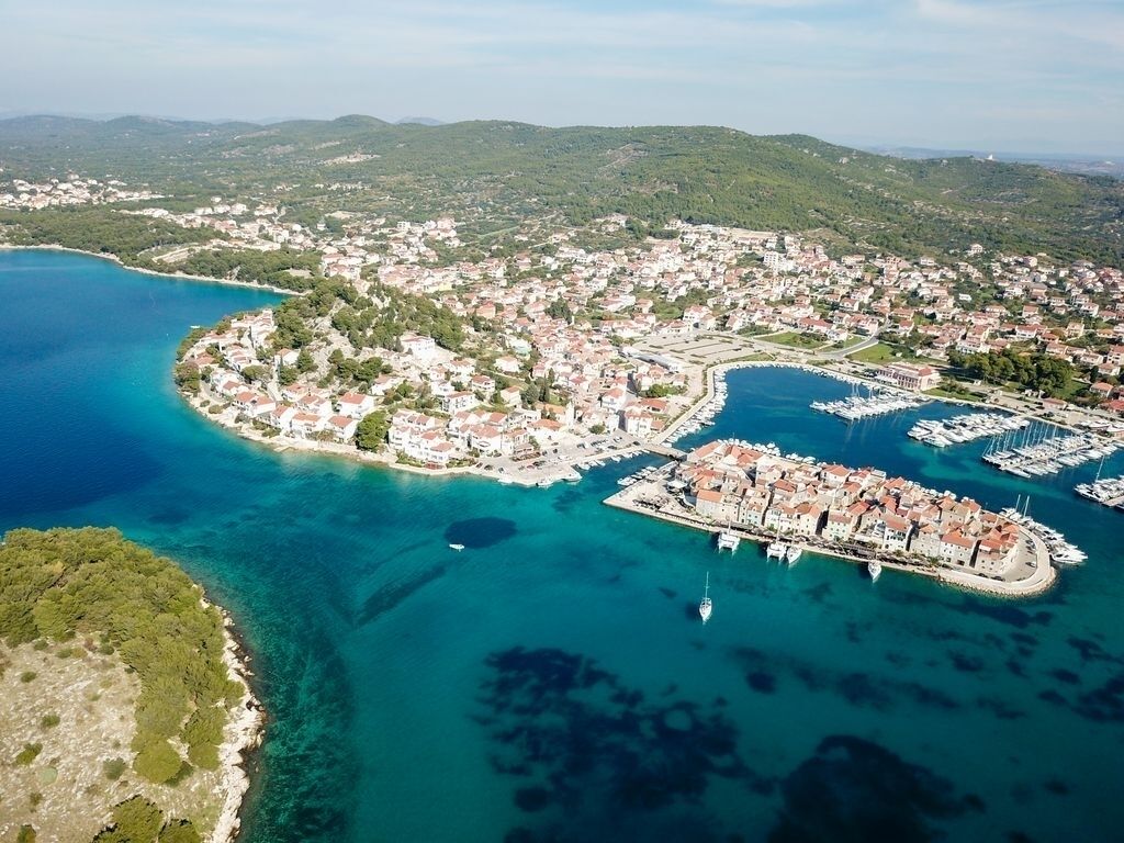 An aerial view of a coastal town with a marina, clear blue waters, and surrounding hills.