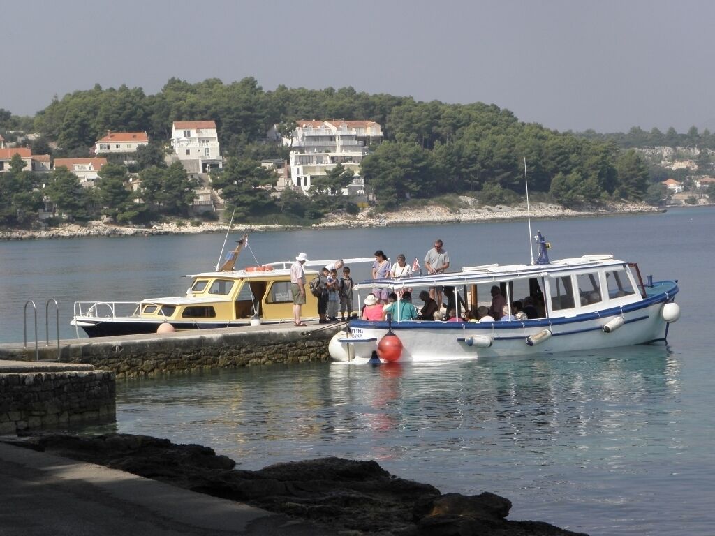 A boat docked at a pier with people on board, a rocky shoreline, and buildings in the background.