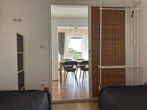 A room with two beds, a sliding door leading to a balcony, a table with chairs, and a view of greenery outside.