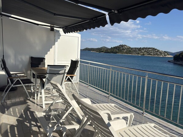 A balcony with white lounge chairs and a table, overlooking a body of water and a hilly landscape.