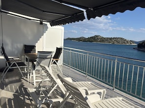 Ein Balkon mit weißen Loungesesseln und einem Tisch, mit Blick auf ein Gewässer und eine hügelige Landschaft.