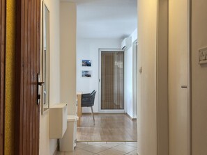 Un couloir avec une porte en bois, un petit coin salon avec un fauteuil, un mur blanc orné de tableaux et une porte vitrée donnant sur un balcon.