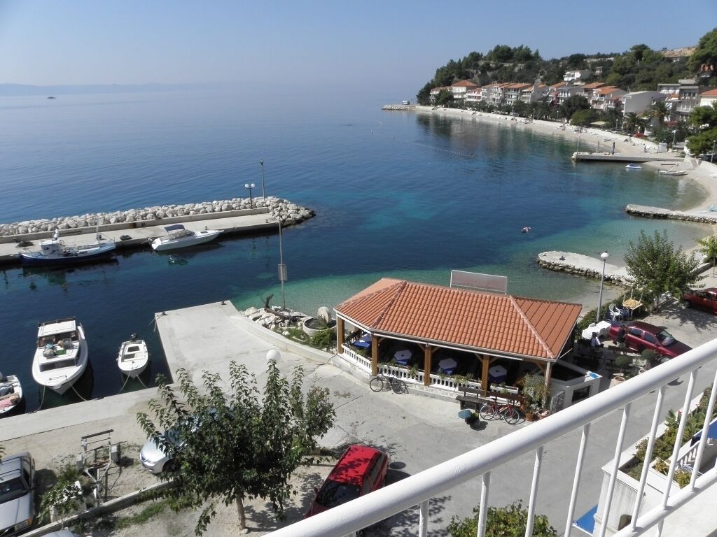 A coastal marina with boats docked, a restaurant with outdoor seating, and a view of the sea and nearby buildings.