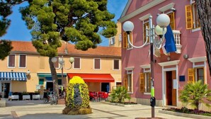 A square with a central fountain, surrounded by buildings with traditional architecture, including a pink building with a balcony and a yellow building with a red awning. - Apartment in Pakoštane with Terrace, Air condition, WIFI, Washing machine (3548-4) (Pakoštane)