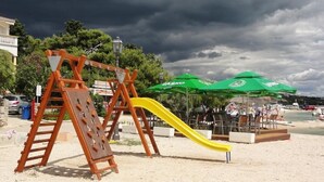 A playground with a yellow slide and a wooden climbing structure, set against a backdrop of a beach and a cloudy sky. - Apartment in Pakoštane with Terrace, Air condition, WIFI, Washing machine (3548-4) (Pakoštane)