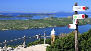 A wooden signpost with directional arrows and distance markers, a man standing on a stone platform, and a scenic view of a lake with islands. - Apartment in Pakoštane with Terrace, Air condition, WIFI, Washing machine (3548-4) (Pakoštane)