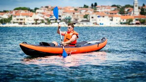 Eine Person paddelt mit einem Kajak auf dem Wasser, im Hintergrund ist eine Stadt zu sehen.