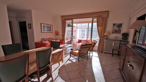 Una sala de estar con comedor, televisor y radiador. Se observa una ventana con cortinas y un cuadro en la pared.